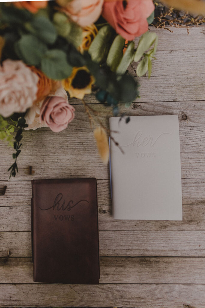 Photo showing the florals and vow books of the bride and groom against a wood background.