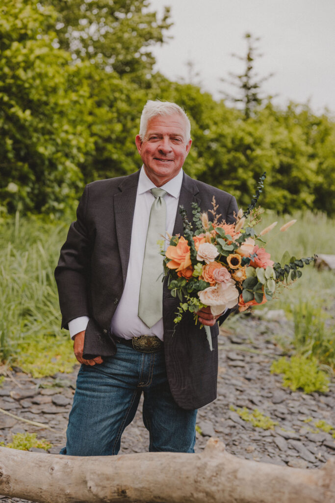 Father of the Bride holds her bouquet before walking down the aisle of the elopement she planned in Seward.