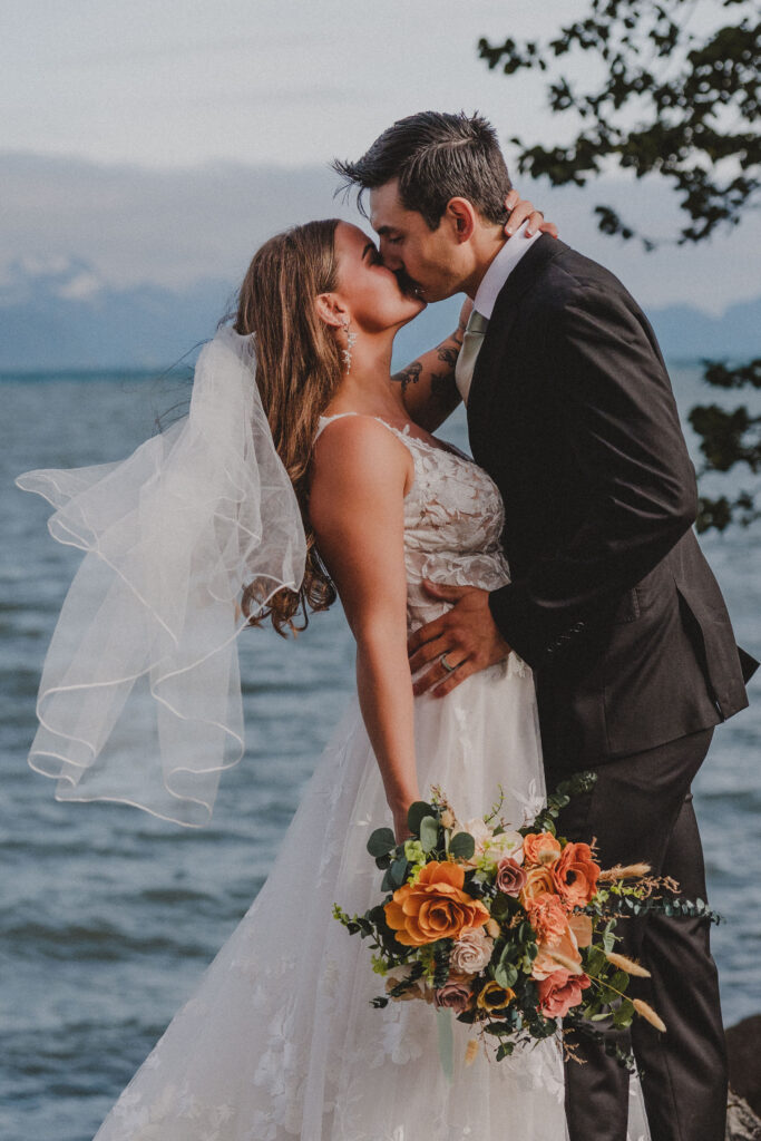 The bride and groom kiss as the bride's veil flutters in the wind after their beach wedding at Lowell Point.